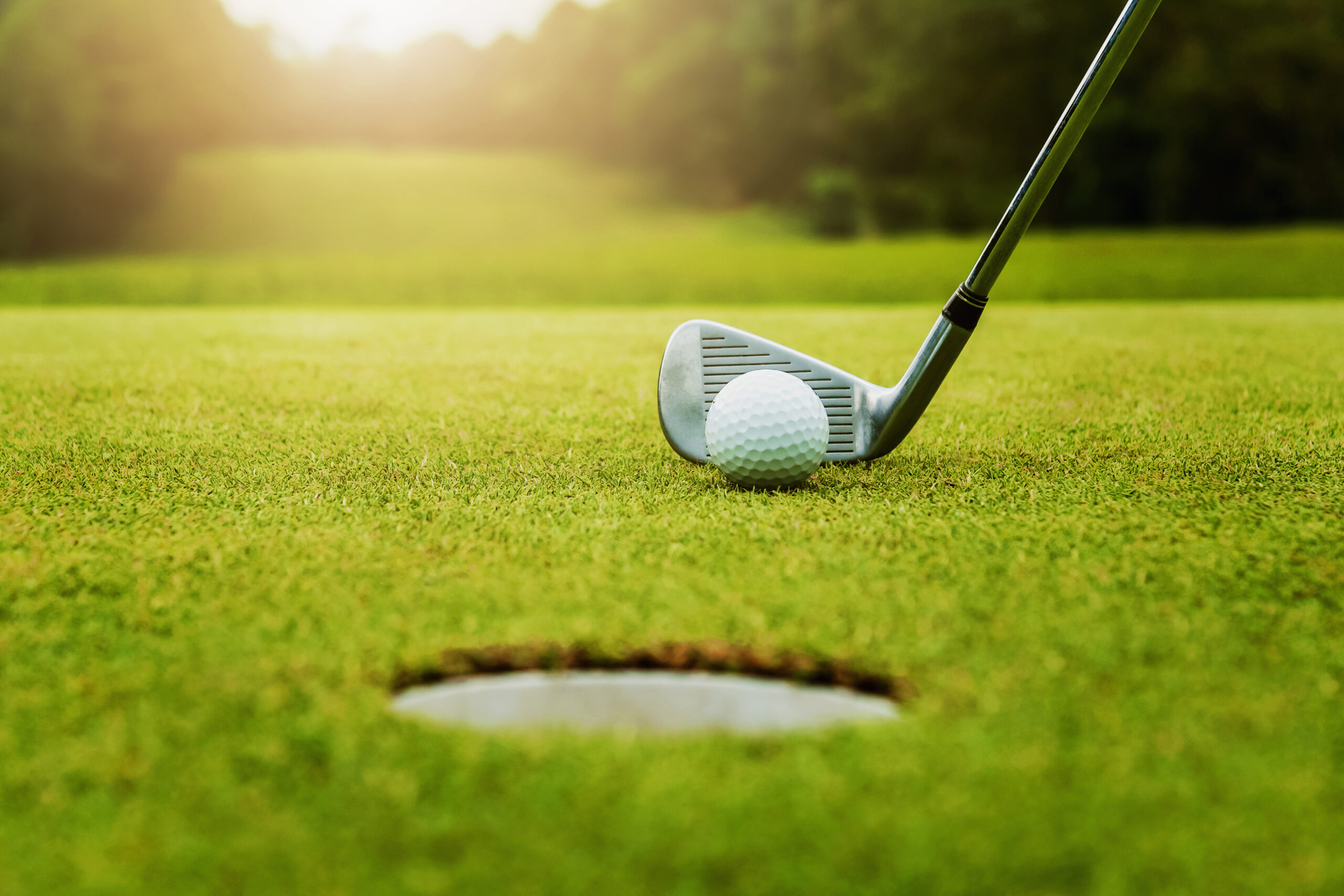 close up golf club and golf ball on green grass with sunrise bac